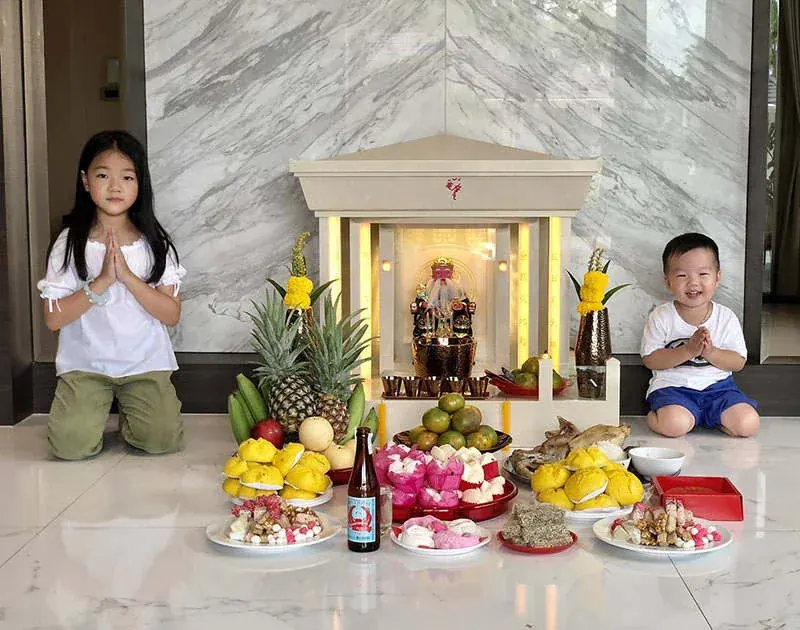 ลูกชายและลูกสาวถ่ายรูปกับ ตี่จู้ หินอ่อน 35 นิ้ว สีเบจน้ำตาลอ่อน ด้านข้างมีเด็กชาย เด็กหญิง กำลังพนมมือไหว้ ด้วยท่าทางยิ้มแย้ม แจ่มใส เป็นภาพที่ดูอบอุ่นยิ่ง
